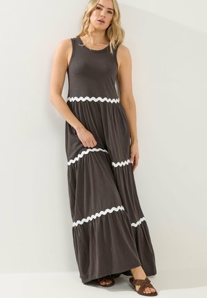 Frau trägt ein ärmelloses, dunkelgraues Maxikleid mit weißen wellenförmigen Streifen, steht vor einer schlichten hellen Wand und hält den Saum des Kleides.