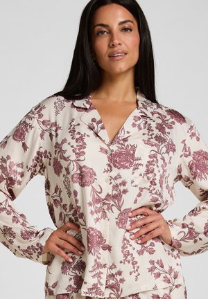 Vrouw die een crèmekleurige blouse draagt met een donkerroze bloemenprint, staand met handen in de zij tegen een effen lichtgrijze achtergrond.