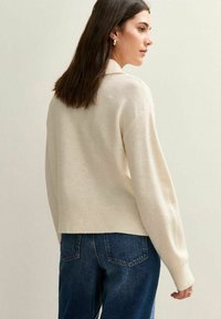 Kobieta z brązowymi włosami do ramion, nosząca złote kolczyki w kształcie kół, kremowy sweter i ciemnoniebieskie dżinsy, stojąca bokiem na jednolitym tle.