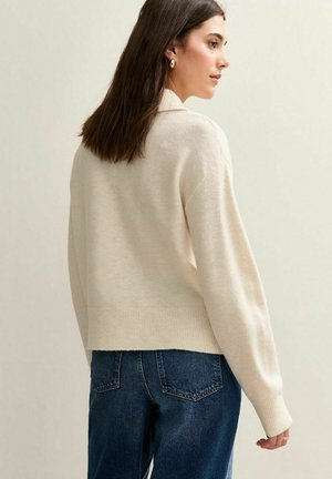Vrouw met lang bruin haar, draagt gouden hoopoorbellen, een crème trui en blauwe jeans, iets naar rechts gedraaid tegen een effen achtergrond.