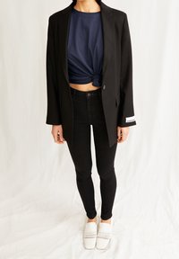 Blazer negro entallado sobre un crop top anudado azul marino, combinado con jeans ajustados negros y zapatos blancos tejidos sin cordones. Diseño simple y moderno.