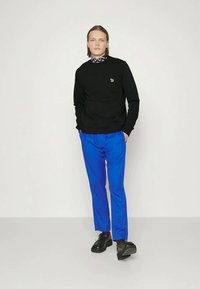 Vállig érő hajú személy fekete pulóverben, kék nadrágban, mintás galléros ingben és fekete cipőben, egy sima fehér háttér előtt áll.