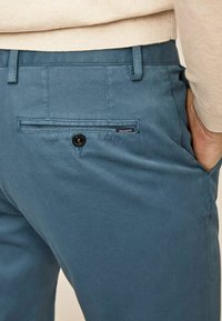 Light blue cotton trousers with a back pocket and black button. Features a stitched logo tag for brand identification.