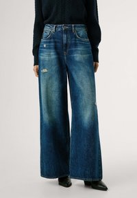 Vaqueros de mezclilla azul con perneras anchas y desgastadas, combinados con una blusa de manga larga negra texturizada y zapatos de tacón negro con punta. 