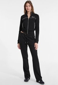 Giacca tuta nera con cerniera e piping bianco, abbinata a pantaloni della tuta coordinati con coulisse regolabile. Entrambi sono realizzati in tessuto morbido.