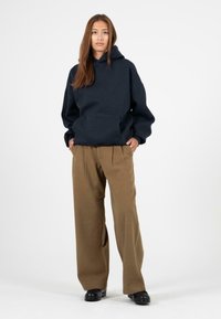 Granatowy bluza z kapturem z przednią kieszenią i luźnym krojem, w połączeniu z brązowymi spodniami o szerokich nogawkach. Model stoi z rękami w kieszeniach.