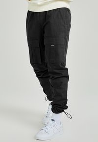 Svarta cargobyxor med stora sidofickor och justerbara ankelslipsar, bärs med vita Nike-sneakers och vita ankelsockor.