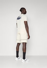 Light beige short-sleeve t-shirt with navy crocodile logo on back, paired with beige shorts with black piping, worn with white sneakers and black socks.