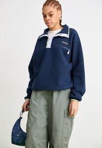 Giovane vestito con giacca in pile blu navy e pantaloni cargo verdi, che tiene in mano una piccola borsa blu, in piedi davanti a uno sfondo bianco semplice.