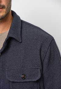 Marineblau strukturierte Jacke mit einem Kragen und einer Brusttasche mit Knopfverschluss. Das weiche Material erhöht den taktilen Reiz.