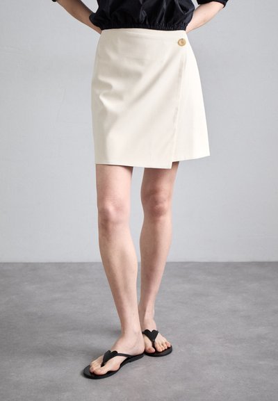 Jupe portefeuille blanche avec un détail de bouton sur le côté, associée à des tongs noires. Le tissu semble lisse avec un design épuré et une coupe ajustée.