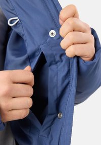 Blå jacka med en ficka med dragkedja; har en metallknapp och elastisk snörning. Tyget verkar slätt och väderbeständigt.