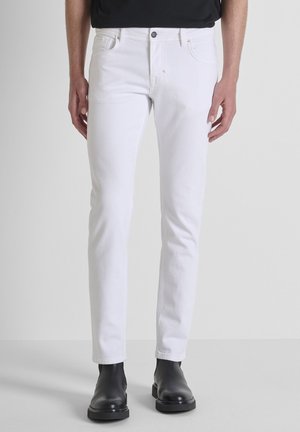 Man draagt slanke witte jeans en zwarte leren enkellaarsjes tegen een effen grijze achtergrond.