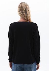 Maglione nero a maniche lunghe con taglio rilassato, ampio scollo e polsini a coste, abbinato a jeans azzurri chiari; texture liscia.