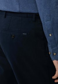 Blaues Texturhemd mit Knopf-Akzenten, kombiniert mit dunkelblauen Hosen, die eine Gesäßtasche und ein kleines Markenetikett haben.