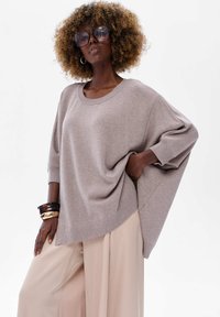 Jasno-beżowy, oversize sweter z prążkowanymi mankietami i ściągaczem. Połączony z szerokimi beżowymi spodniami oraz nałożonymi bransoletkami na jednym nadgarstku.