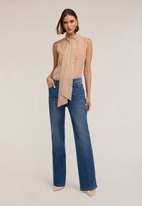 Beżowa bezrękawnik z detalem w postaci węzła, ozdobiona małymi koralikami, w połączeniu z szerokimi jeansami z niebieskiego denimu.