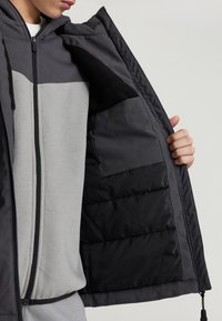 Kurtka z kapturem w ciemnoszarym kolorze z jaśniejszym szarym panelem, z ocieplanym wnętrzem i czarnym polarem w środku, a także z kieszenią wewnętrzną.