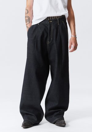 Persona que lleva puestos unos jeans de mezclilla oscuros de pierna ancha, una camisa blanca, zapatos negros, con un tatuaje visible en el brazo y anillos y pulsera de plata.