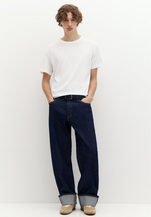 Jeune homme aux cheveux bouclés portant un t-shirt blanc, un jean large retroussé foncé, des baskets beige et les mains dans les poches sur un fond blanc.