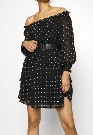 Kobieta w czarnej sukience z odkrytymi ramionami w białe kropki, z przezroczystymi długimi rękawami, warstwową spódnicą i szerokim czarnym paskiem z prostokątną klamrą.