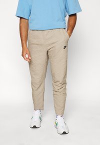 Beige, strukturerade joggers med elastisk midja och en svart Nike-logga. Matchade med en ljusblå skjorta och vita sneakers.