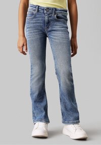 Světle modré džíny s rozšířenými nohavicemi, pětikapsovým designem, jemným prošoupáním a kovovým zapínáním na knoflík. Nošeny s bílými teniskami.