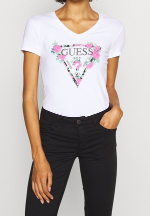 Woman wearing a white V-neck T-shirt with a pink floral GUESS logo and black pants, standing with one hand in pocket.