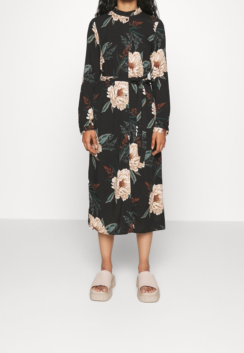 Robe noire à fleurs avec manches longues, taille ceinturée et devant boutonné. Motif de pivoines beiges et feuilles vertes. Portée avec des sandales de couleur claire.