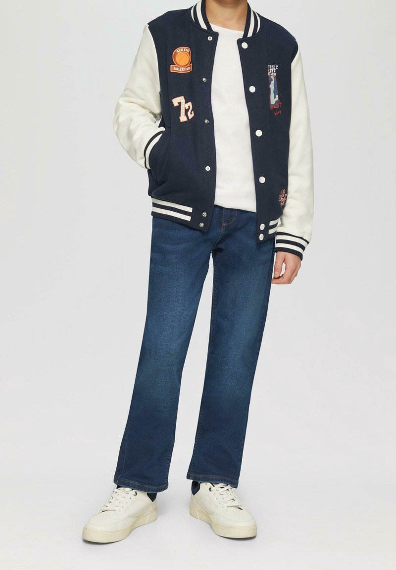 Veste universitaire bleu marine et crème avec écussons brodés, poignets rayés et boutons-pression classiques, associée à un jean bleu foncé et des baskets blanches.