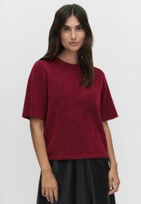 Czerwony sweter z krótkim rękawem, wykonany z miękkiego, puszystego materiału, z okrągłym dekoltem i luźnym krojem, w zestawie z czarną plisowaną spódnicą.