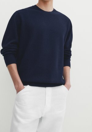 Persoon die een textuur marineblauwe trui met lange mouwen draagt, met handen in de zakken van een witte broek tegen een effen achtergrond.