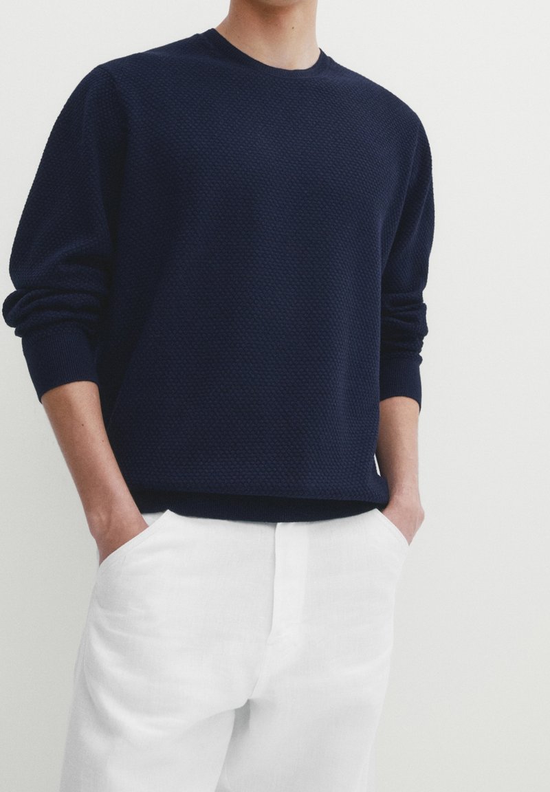 Personne portant un pull à manches longues texturé bleu marine avec les mains dans les poches d'un pantalon blanc, sur fond uni.