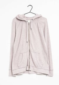 Hoodie zippé rose clair avec cordons et poches avant, suspendu à un cintre noir sur fond blanc.