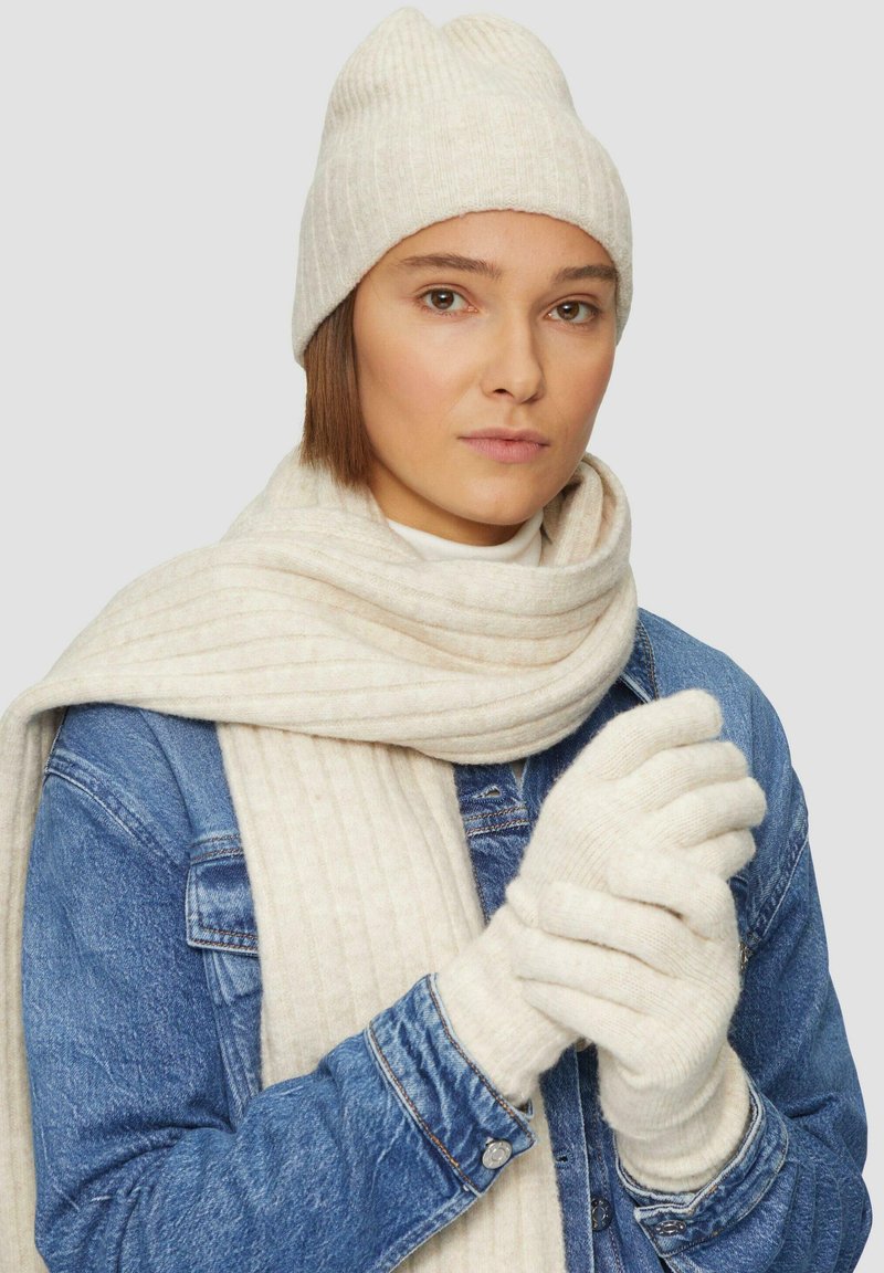 Gebreide beanie, sjaal en handschoenen in zachte crèmekleur; sjaal heeft verticale ribbels, handschoenen hebben een gestructureerd oppervlak; eronder een spijkerjack.