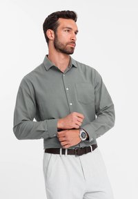 Chemise à manches longues vert clair avec un design à boutonnière et une poche poitrine. Présente des boutons blancs et une coupe décontractée. Associée à un pantalon beige.