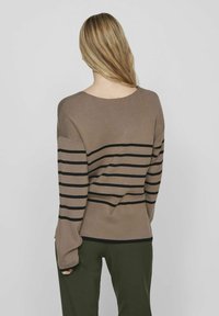 Maglione marrone a righe con strisce orizzontali nere, texture in maglia, vestibilità oversize, scollo rotondo e maniche lunghe, abbinato a pantaloni verdi.