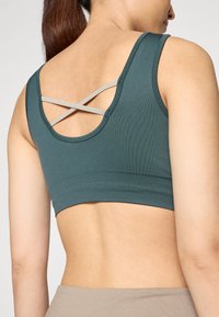 Sujeción deportiva en tono verde azulado con textura acanalada y espalda redondeada. Presenta tirantes cruzados en gris claro para mayor soporte.