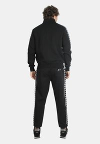Uomo che indossa un completo sportivo nero con strisce bianche con logo su maniche e pantaloni, in piedi di spalle su uno sfondo bianco uniforme.