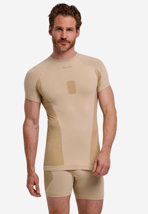 Man met krullend haar die een beige sportshirt met korte mouwen en bijpassende shorts met gestructureerde paneeldetails draagt, tegen een witte achtergrond.
