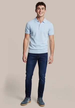 Jonge man glimlachend, staand, draagt een lichtblauw poloshirt, donkerblauwe spijkerbroek en blauwe sneakers met rubberen zolen tegen een effen achtergrond.