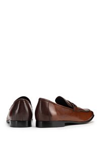Mocassins en cuir marron avec une texture lisse, une pointe arrondie et une couture subtile. Présentent une sangle décorative sur le dessus. Semelle noire.