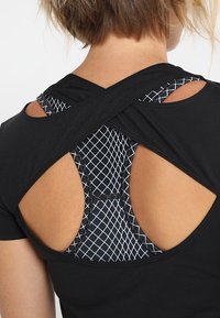 Svart atletisk topp med korsad ryggdesign som avslöjar en lager-på-lager svart och vit sportbehå med rutmönster under. Slät textur.