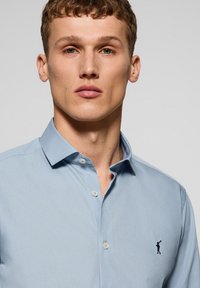 Himmelblaues, knopfbares Hemd mit einem Spread-Kragen, das ein Logo auf der linken Brust aufweist. Gefertigt aus einem glatten, gewebten Stoff mit einer subtilen Textur.