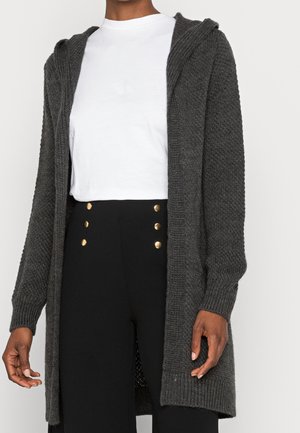 Persona che indossa un cardigan aperto lavorato a maglia di colore grigio scuro, una maglietta bianca con scollo rotondo e pantaloni neri a vita alta con bottoni dorati.