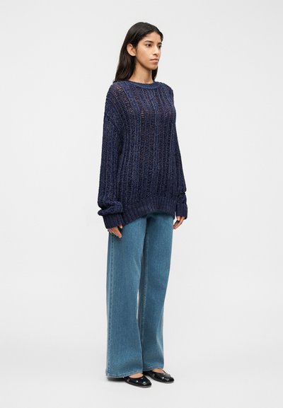 Pull en maille bleu marine à manches larges, présentant un design ample et texturé. Associé à un jean bleu clair à coupe décontractée et des chaussures noires sans lacets.