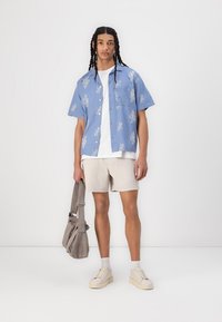 Chemise bleu clair à manches courtes avec motif floral blanc, t-shirt blanc, shorts beiges, baskets blanches et sac à dos gris.