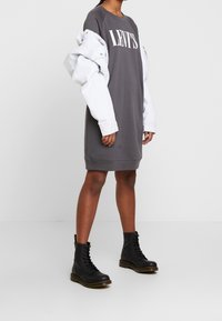 Šedé šaty v podobě mikiny s nápisem "LEVI'S", vrstvené pod světlou modrou denimovou bundou. Spárováno s černými šněrovacími botami. Jednoduchý design, ležérní styl.