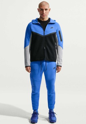 Mand iført blå, sort og grå Nike lynlåstrøje, matchende blå Nike joggingbukser og blå træningssko, der står mod fronten på en ensfarvet baggrund.