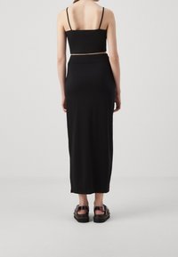 Schwarzes Cropped Top mit dünnen Trägern, kombiniert mit einem schwarzen Maxirock. Glatte Textur, figurbetontes Design, getragen mit klobigen schwarzen Sandalen.